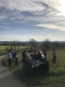 foto med børn og vokse i samtale udendørs med grønne marker og blå himmel i baggrunden